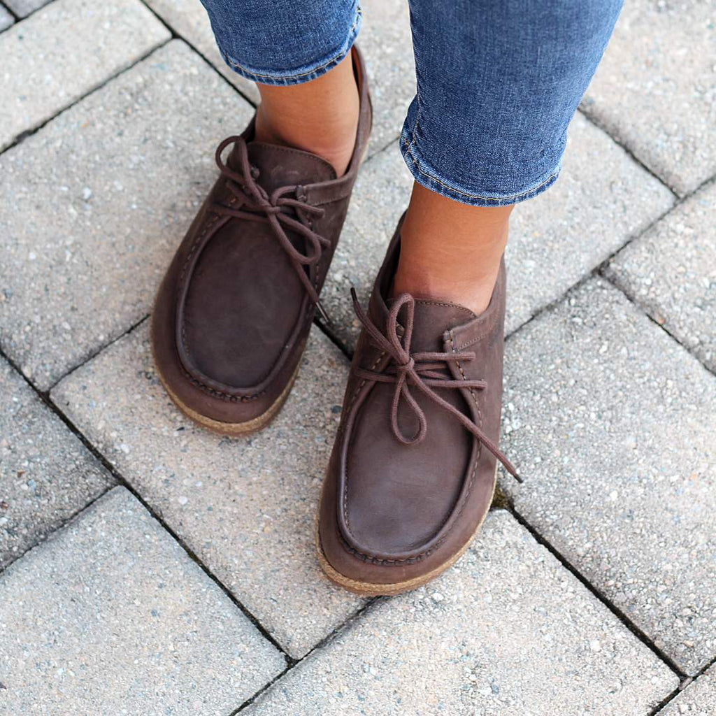 A person wearing blue jeans stands on a stone pavement, sporting the Women's 'Mainz' Moc Shoes. These shoes boast oiled leather uppers that lend a rustic elegance, while the UltraComfort footbed ensures all-day support.