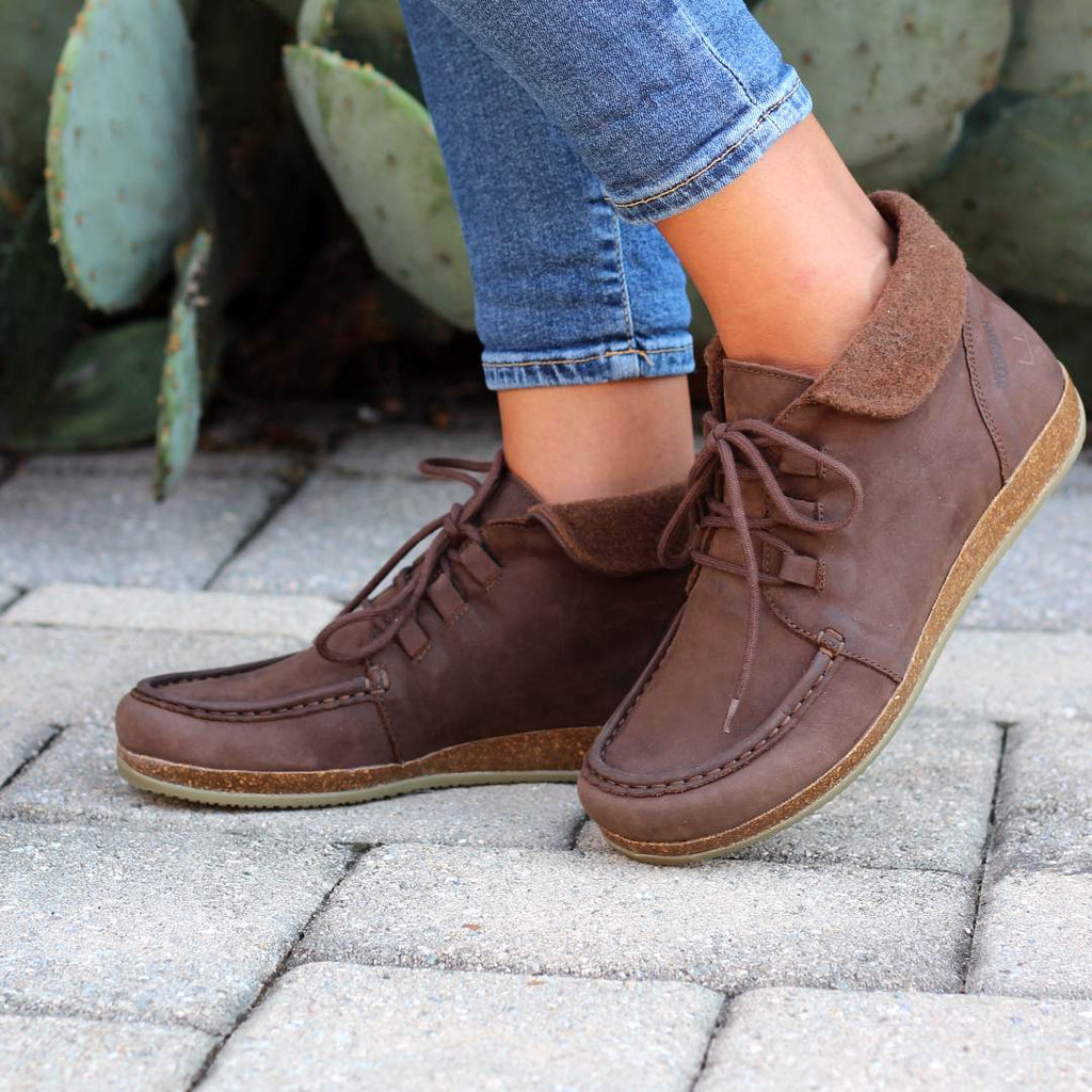 Close-up of a person wearing Women's 'Baden' Boots with jeans, standing on a stone pavement with large green plants in the background.