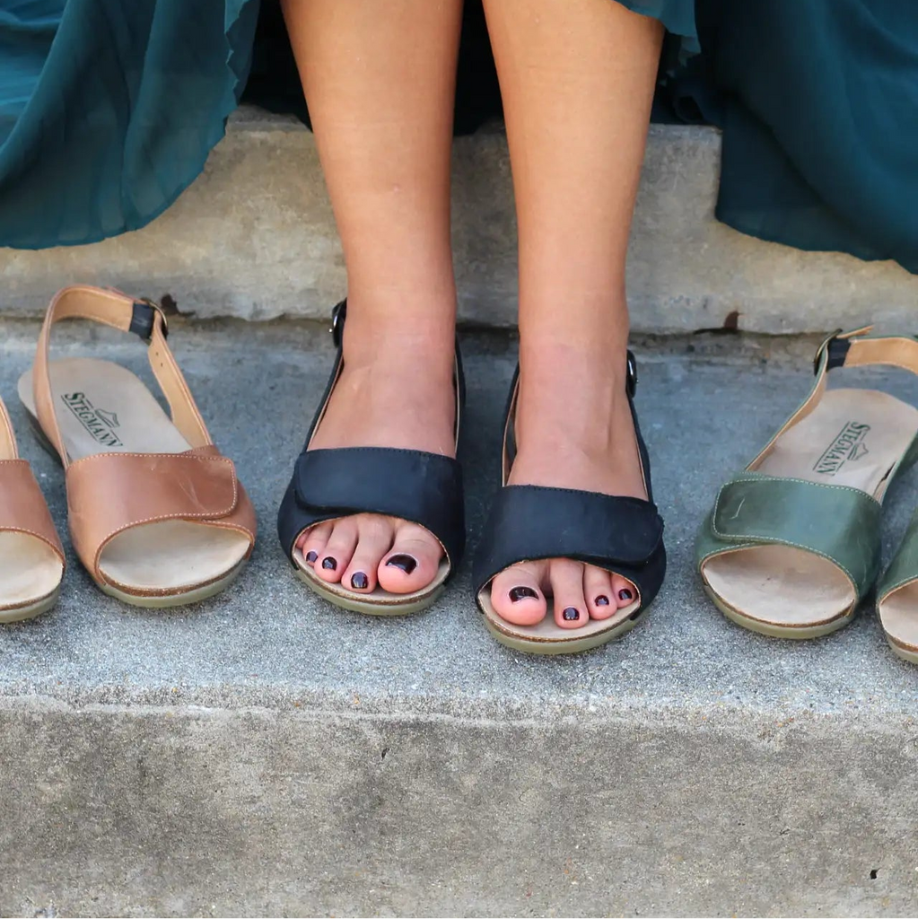 A person sits on steps wearing black Women's 'Clara' Slingback Sandals with padded arch support, flanked by tan cushioned footbed sandals on the left and green supportive sandals on the right.