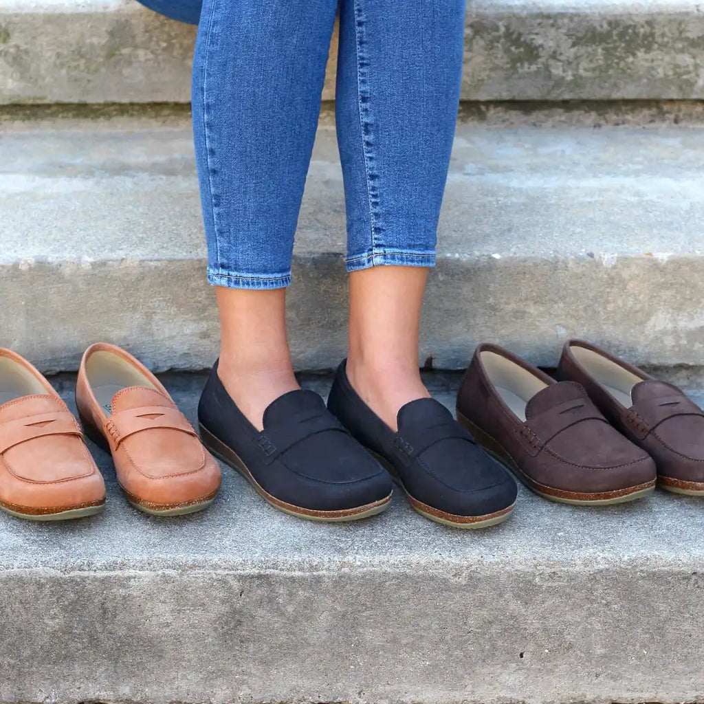 A person in blue jeans and Women's Victoria Loafers with shock-absorbing midsoles sits on steps, surrounded by four pairs of loafers in tan, black, and brown.