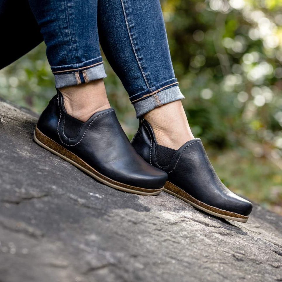 A person wearing Women's Lieben Leather Chelsea Boots with vegetable-tanned leather and rolled-up jeans sits on a rock in an outdoor setting.