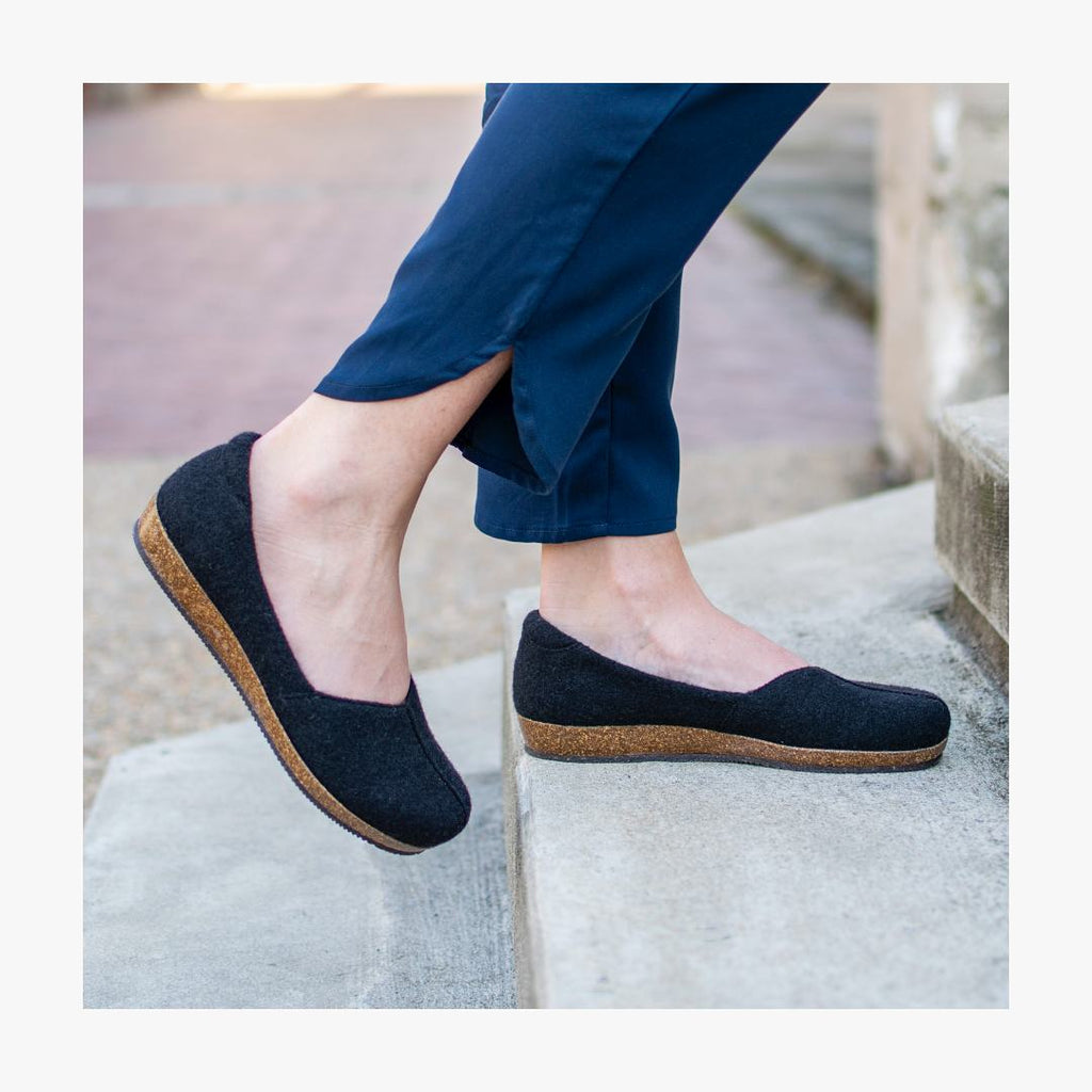 Person stepping on a concrete stair wearing Women's Liesl Wool Skimmer shoes, featuring black slip-on style with cork and anatomically shaped support soles, paired with navy blue pants.