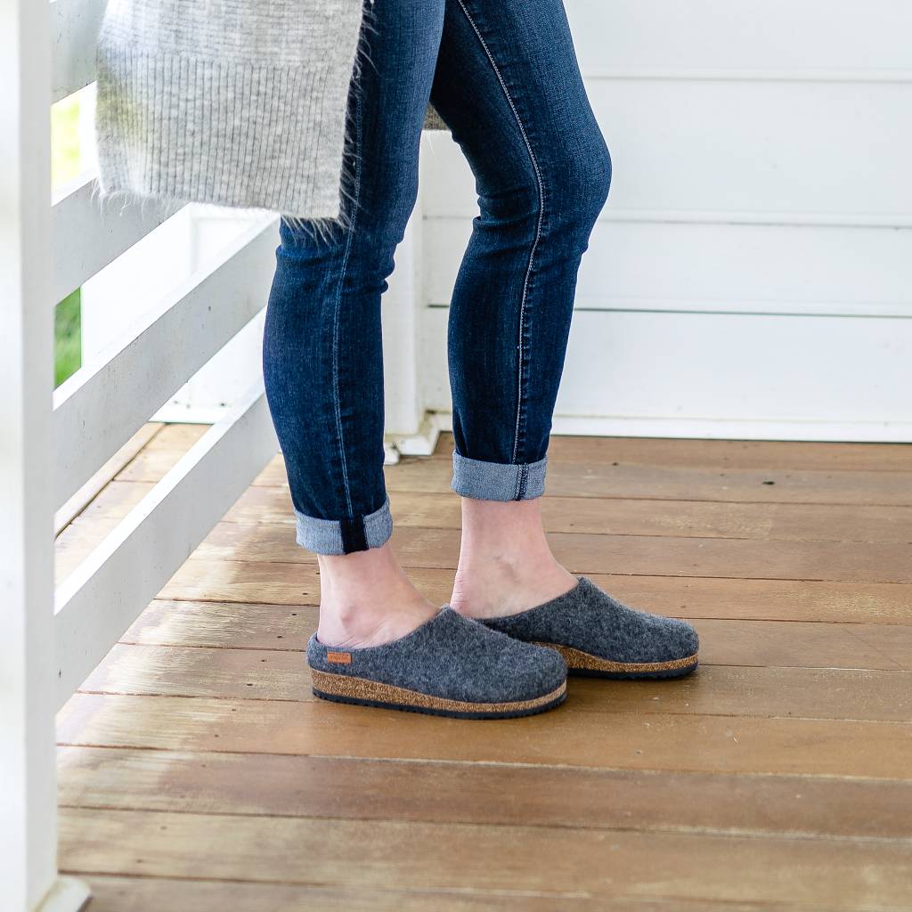 A person in rolled-up jeans and Women's Essenz Wool Clog slippers stands on a wooden porch, leaning against a white railing.