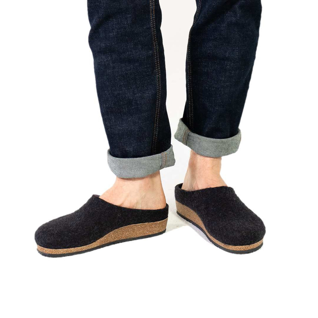 A person wearing dark blue jeans and a pair of Men's Essenz Wool Clog with a cozy Merino wool blend upper and black slip-on design with brown soles is standing on a white background.